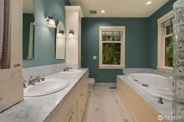 Five-piece primary bathroom has double vanity with Carrera marble counters and floor trim, heated tile floor, luxury shower system and drop-in jetted tub with tile surround