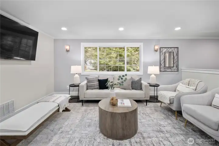 The living room carries a calm, welcoming energy centered around a multi paned window that brings the surrounding evergreens gently into view. Modern sconces with subtle Art Deco influence frame the window in quiet symmetry, their clean vertical lines adding structure while casting a soft ambient glow in the evenings. Crown molding traces the ceiling, lending a refined architectural finish that subtly elevates the space.  Soft light gray tones surround the room, complemented by crisp white trim and the golden beige hues just beyond on the upper level. This warm cool contrast feels fresh yet timeless, creating a palette that works beautifully with nearly any style while maintaining a natural flow throughout the home. Natural light filters in easily, enhancing the layered tones and giving the room an inviting, grounded warmth. You'll find new lighting, new matching hardware finishes, new outlet covers and light switches, fresh paint, and new flooring throughout the entire home.