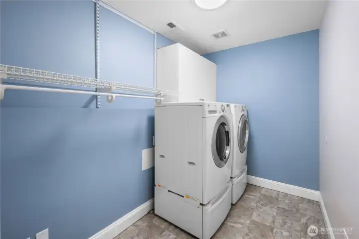 The lower level laundry room is generously sized, giving you plenty of room to move around and keep things organized. A full front-load washer and matching dryer sit side by side, ready for everyday loads or larger laundry days.  Open shelving and hanging space stretch along the wall, offering room for baskets, linens, and sorting stations, while still leaving floor space for a folding table or additional storage if you choose. Bright paint tones keep the space feeling cheerful and clean, turning a practical task into a pleasant part of the day.