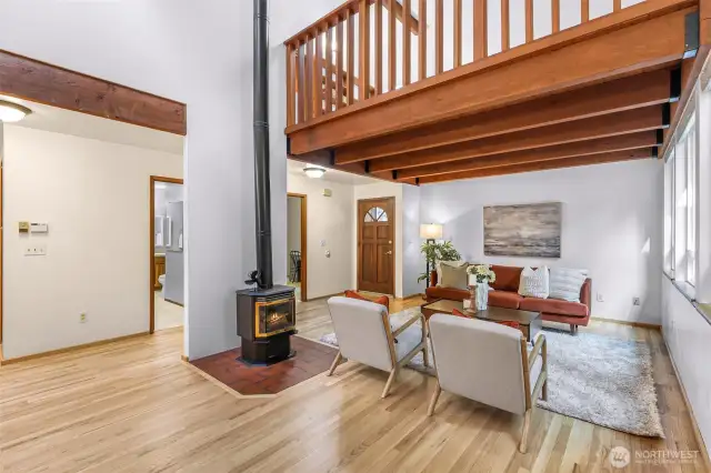 Anchored by the central wood stove, this view of the living area captures the cozy layout beneath exposed beams and the warmth of the upstairs mezzanine.