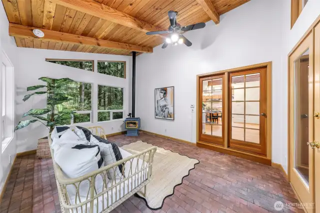 This sunroom glows with character—exposed beam ceiling, brick floors, and oversized windows that frame the surrounding evergreens. A freestanding gas stove brings warmth and style.