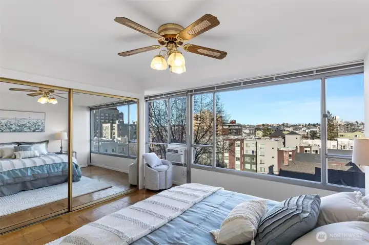 The bedroom keeps the same bright, open feel with expansive windows, a generous mirrored closet that amplifies the space, and a window A/C unit for warm days.