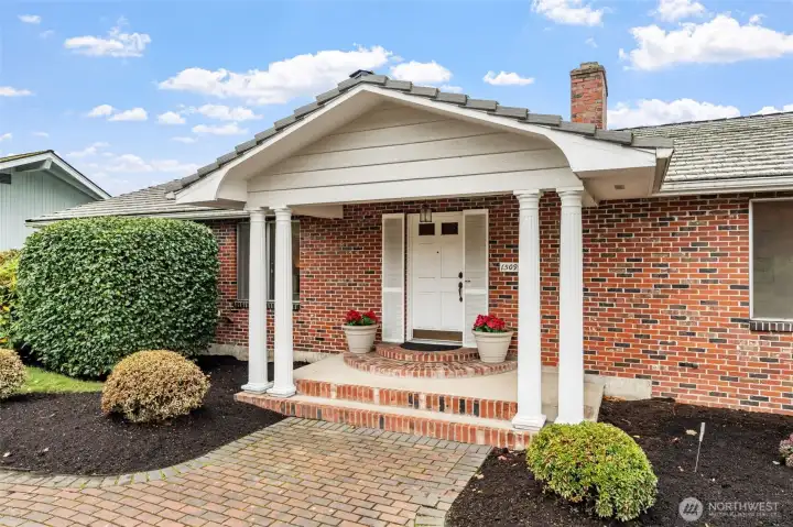 Covered front porch with classic columns offers sheltered entry for guests in all weather. Brick steps are framed by tidy landscaping and a paver walkway.