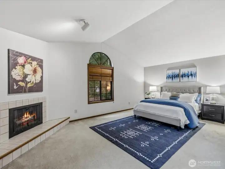Expansive upper primary suite w vaulted ceilings, a cozy fireplace, & a signature arched window framing lush Pacific Northwest greenery