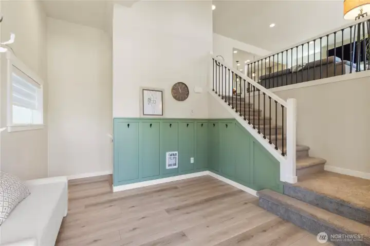 Main landing highlights wide-plank luxury vinyl plank flooring and 2025 custom board-and-batten style green paneling with integrated jacket hooks. Plush carpeted stairs lead up. (Furniture does not convey) Main landing highlights wide-plank luxury vinyl plank flooring and 2025 custom board-and-batten style green paneling with integrated jacket hooks. Plush carpeted stairs lead up. (Furniture does not convey)