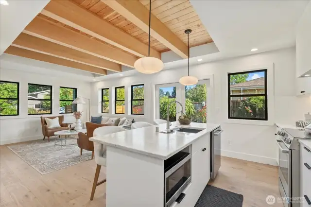 Bright, spacious open concept living, kitchen, dining area with custom exposed-beam ceiling, upgraded black Milgard windows, wide-plank hardwood flooring, and natural-stone countertops.