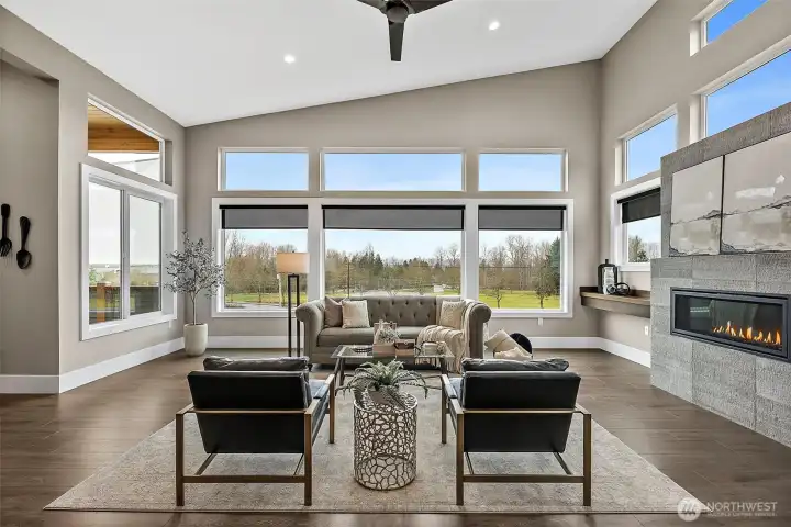 The living room features tall windows that frame open views while filling the space with natural light. A modern fireplace and open concept layout create a bright and welcoming living area designed for both entertaining and everyday comfort.