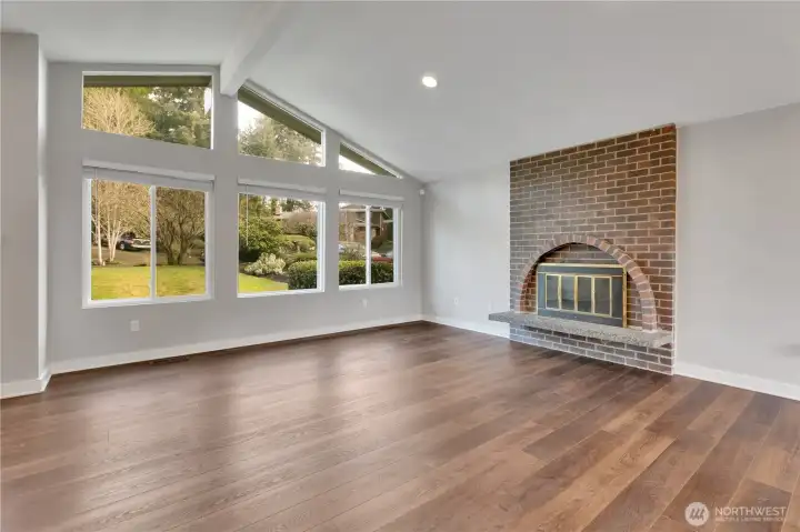 Spacious living room featuring large windows, brick fireplace, and vaulted ceiling.