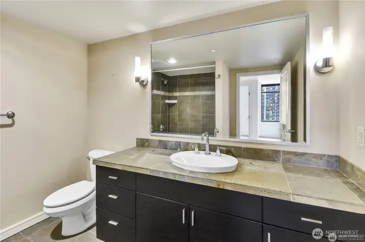 The bathroom features a tiled shower-tub combination with earthy stone finishes, complementing the vanity design.