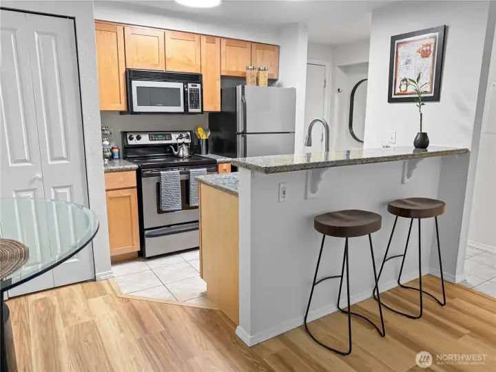 Another view of the kitchen and eat-in bar, this time as viewed from the edge of the dining room / beginning of the living room. The kitchen boasts granite counters and stainless steel appliances. The stacked washer and dryer and water heater are in the closet on the left side of the photo.