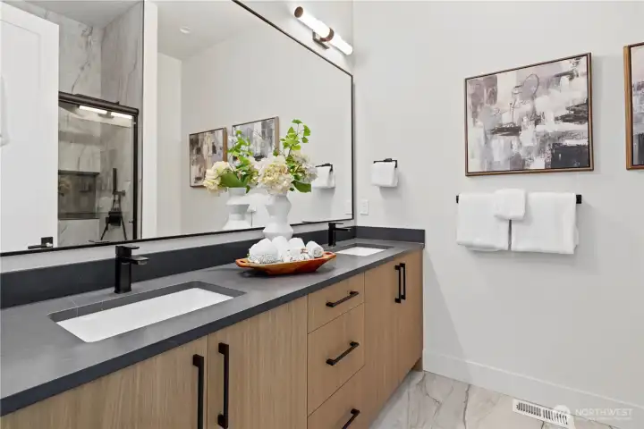 Sleek main-floor ensuite featuring double vanity with leathered quartz counters, matte black fixtures, framed mirror, and heated tile floors.