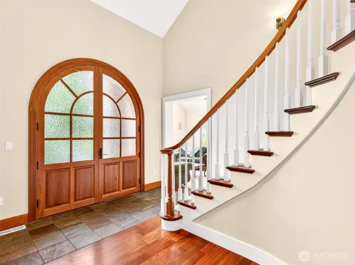 The arched wood-and-glass entry door bathes the foyer in natural light, framed by vaulted ceilings and the elegant sweep of the curved staircase. A grand yet inviting introduction to the craftsmanship that defines this home.