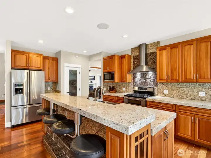 Heavy-duty built-in bar stools tuck neatly under the expansive island, offering casual seating for morning coffee or homework hour. Stainless appliances, double ovens, and a tiled backsplash complete this hardworking gourmet space.