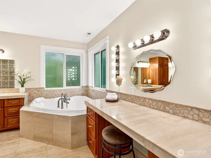 A jetted corner soaking tub sits beneath corner windows, paired with a tile-topped vanity station perfect for getting ready in style. Spa-quality details turn everyday routines into a daily indulgence.