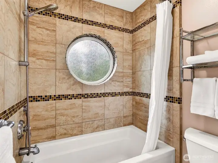 A full tub-shower combo features travertine-style tile, mosaic accent bands, and a charming porthole window that floods the space with natural light. Stylish and timeless for everyday use.