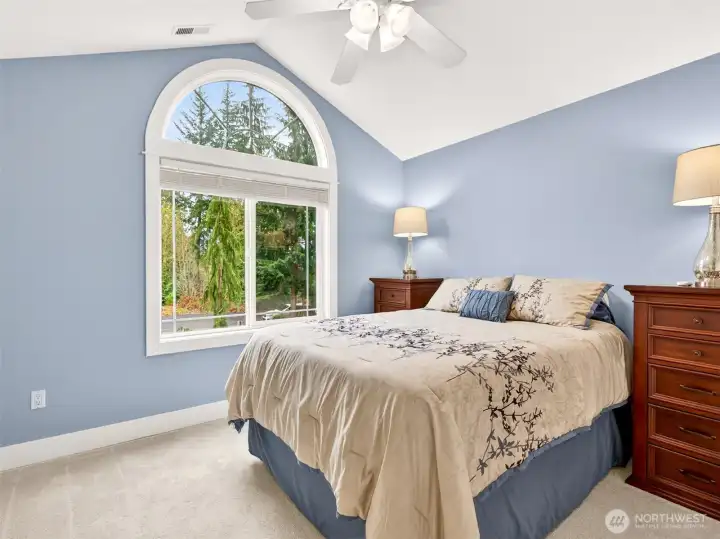 A serene upstairs bedroom features vaulted ceilings, an arched picture window, and treetop views that invite restful sleep. A peaceful retreat for family or guests.