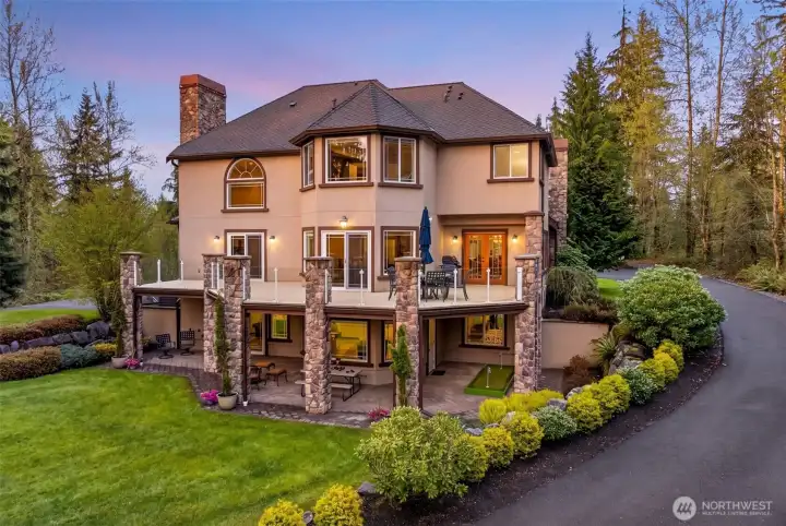 Twilight reveals the home's stunning multi-level rear elevation, with glowing windows, layered patios, and architectural stonework cascading down to the lush grounds. A storybook setting you'll be proud to call home.