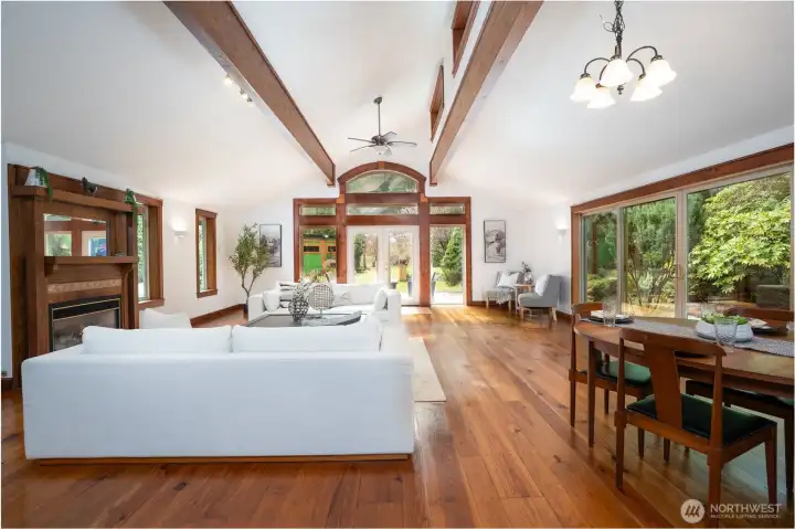 Stunning great room with soaring vaulted ceilings, exposed wood beams, and expansive windows that bring in natural light and serene outdoor views. The open-concept layout seamlessly connects the living and dining areas, centered around a cozy fireplace and framed by rich wood finishes throughout. Large sliding doors provide effortless indoor-outdoor living, creating an inviting space perfect for relaxing or entertaining.