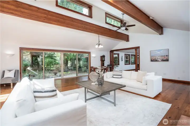 Inviting living space with soaring vaulted ceilings, exposed wood beams, and expansive windows that flood the room with natural light. The open layout is anchored by warm wood flooring and a cozy fireplace, while large sliding glass doors showcase peaceful outdoor views and create a seamless connection to the surrounding landscape, perfect for both relaxing and entertaining.