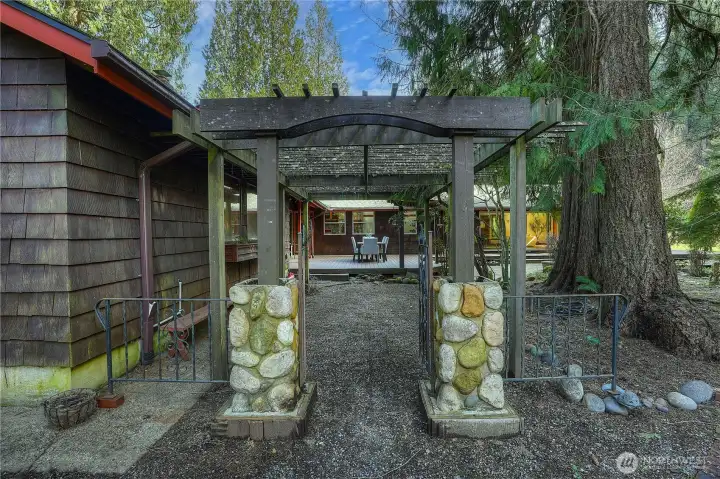 Inviting garden entry with rustic stone pillars and a wood pergola creates a warm transition into the outdoor living space. The pathway leads to a private deck ideal for dining and entertaining, surrounded by mature trees and natural landscaping.
