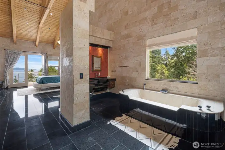 Open primary bath includes a large soaking tub, stone finishes, black tile flooring, and views toward the surrounding evergreens.