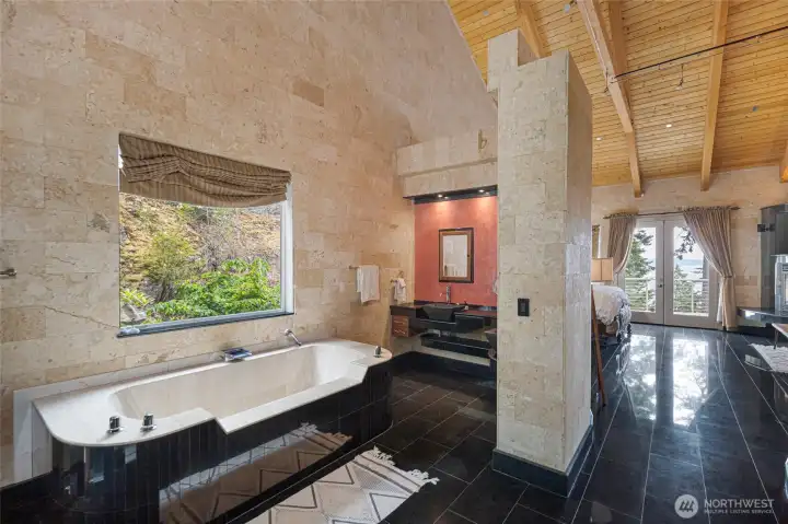 The second primary bath features a large soaking tub, stone walls, dark tile flooring, and an open layout adjoining the bedroom.