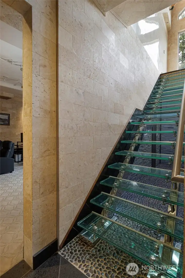 A striking glass staircase with metal railings, stone walls, and skylight illumination connects the home’s main and lower levels.