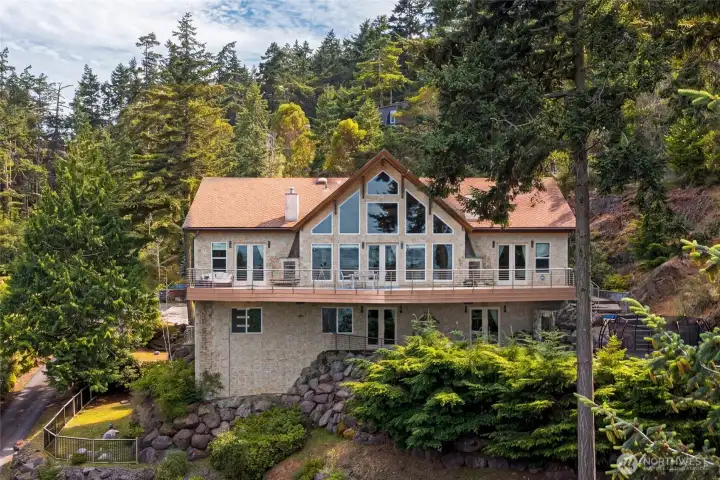 Exterior view showcases the home’s dramatic window wall, broad entertaining deck, lower patio, and custom architectural design.