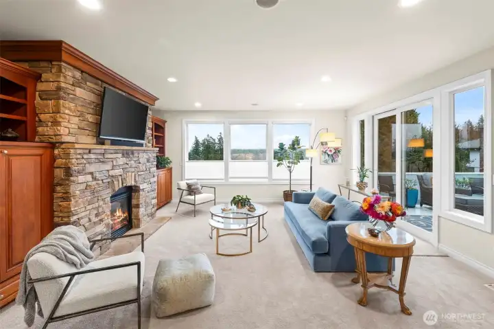 Spacious living room with stone fireplace, custom built-ins, and large windows overlooking Puget Sound and evergreens.