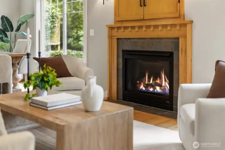A warm, grounded living space anchored by a beautifully crafted fireplace—where natural wood tones, soft light, and curated simplicity create an effortless sense of comfort. Framed by oversized windows and lush greenery beyond, the room invites quiet mornings, intimate evenings, and a seamless connection to the landscape that defines Bainbridge living.