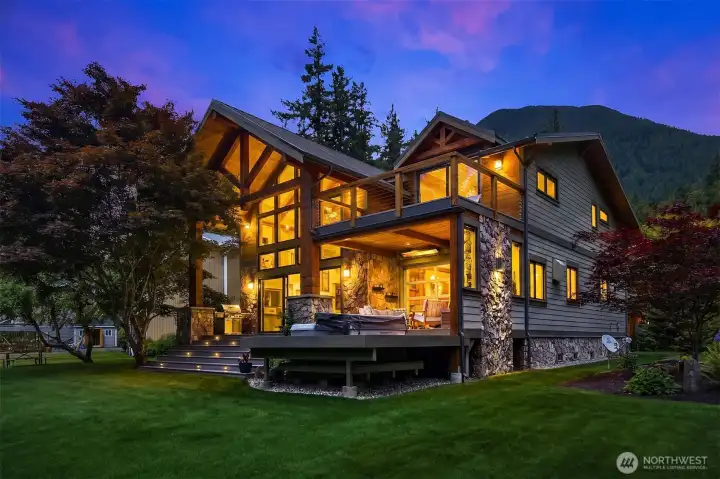 The riverfront exterior glows at twilight, emphasizing the architectural building accent lighting, the primary suite’s private 16'x14' waterproof upper deck, and the elite, year-round covered outdoor patio space overlooking the water.