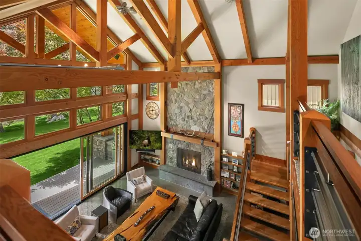 A bird's-eye view from the upper loft highlights the dramatic structural volume, intricate timber framing, and soaring stone hearth. Expansive walls of glass flood the open-concept floor plan with natural light and frame the estate’s private park-like setting.