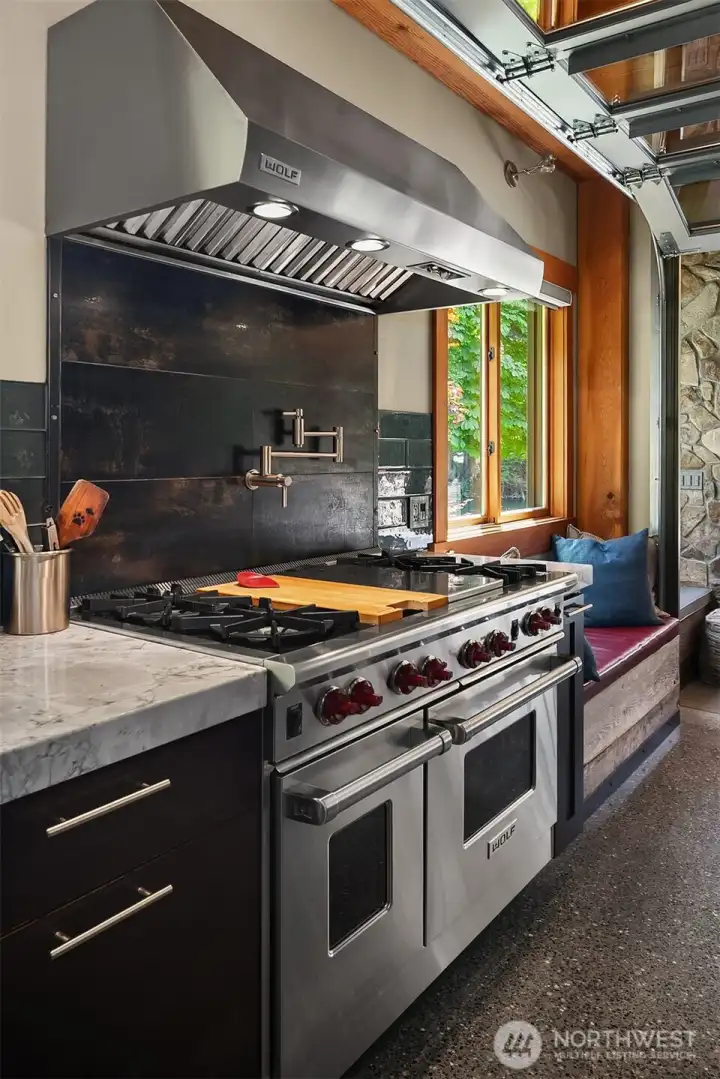 Commercial-grade culinary appointments featuring a premium 48-inch Wolf double-oven gas range with signature red knobs, a custom distressed-metal backsplash, high-capacity stainless hood, and a convenient wall-mounted pot filler.