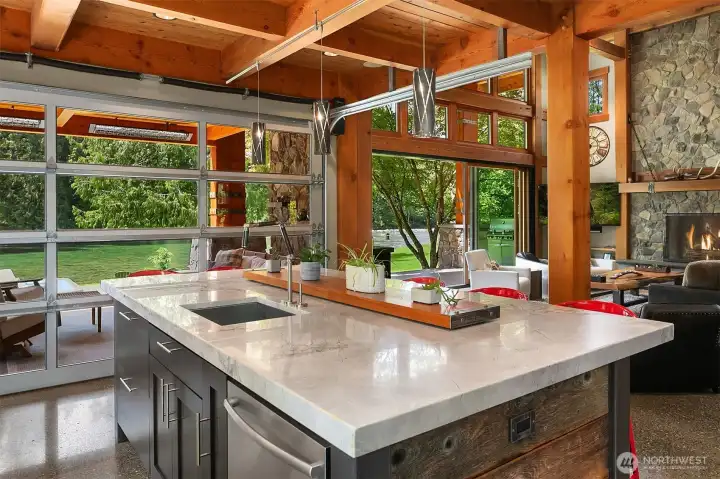 Culinary artistry meets modern industrial design. The gourmet kitchen features a massive stone-topped entertaining island, heated polished concrete floors, and a clear-glass roll-up garage door that opens completely to the riverfront patio.