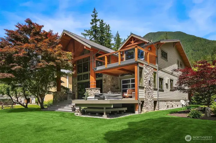 Striking rear elevation showcasing the home's masterfully executed modern Pacific Northwest architecture. Nestled against a dramatic mountain backdrop, the estate features multi-level decks, a recessed spa, and a sprawling lawn rolling down to the water.