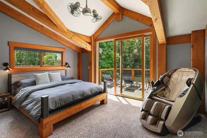 The sanctuary-like primary suite boasts soaring vaulted ceilings, exposed timber frame construction, and large sliding glass doors that step out directly onto a private riverfront deck.