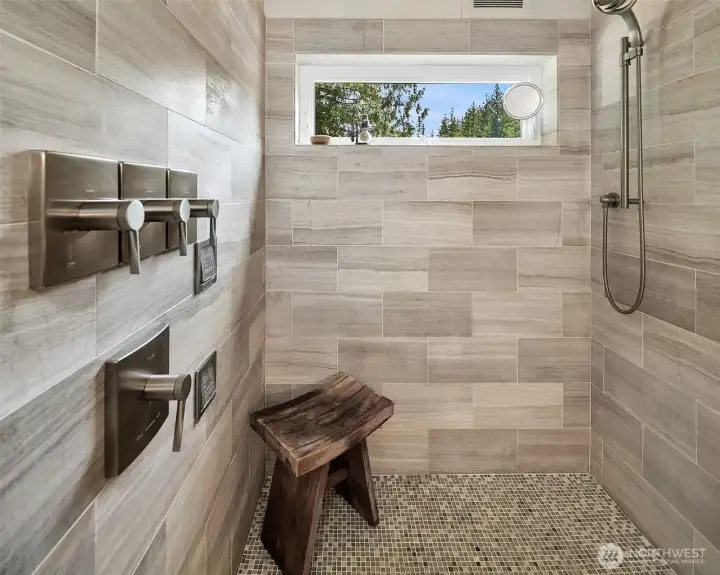 A luxurious walk-in shower lined with premium large-format tile. Fully equipped with an advanced multi-valve shower system, integrated body sprays, a handheld wand, and a high-placed privacy window framing forest views.