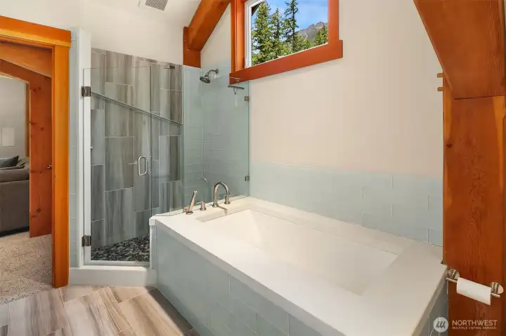 The beautifully appointed upstairs full bath features a deep standalone soaking tub wrapped in soft-blue subway tile, a separate glass-enclosed walk-in tile shower, and contemporary wood-look tile floors.