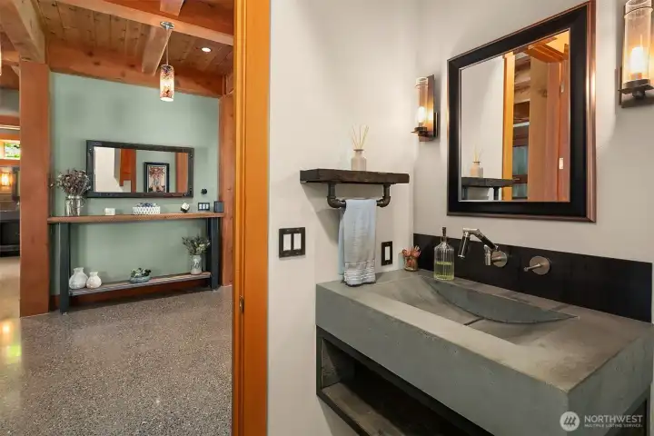 A striking downstairs half bath seamlessly blending industrial elements with modern design. Features a custom-cast ramped concrete sink, wall-mounted matte hardware, a pipe-style towel rack, and rich timber trim.