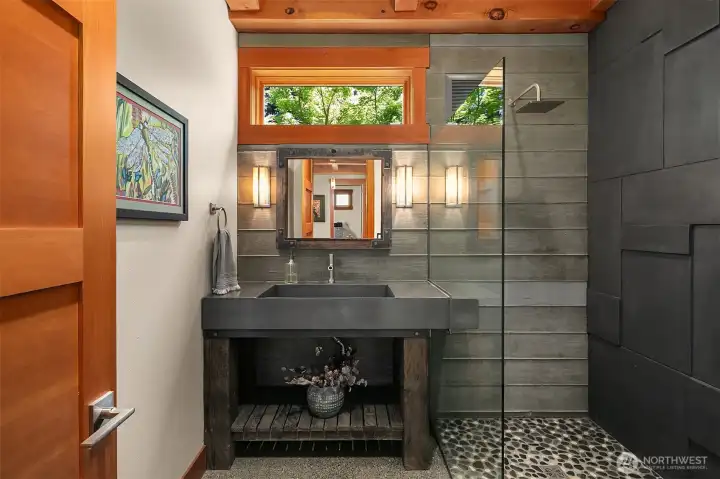 The design-forward downstairs 3/4 bath showcases an architectural, geometric tile accent wall in matte charcoal, a custom concrete trough sink on a heavy timber base, and a seamless glass shower with a natural pebble floor.