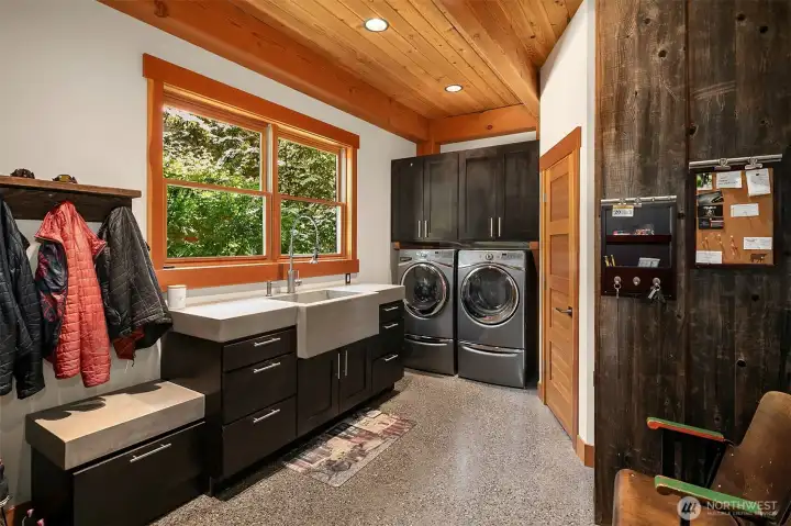 A high-end laundry and mudroom that combines utility with industrial style. Features a custom concrete apron-front utility sink, extensive dark-slab cabinetry, premium front-load laundry appliances, and a reclaimed wood accent wall.