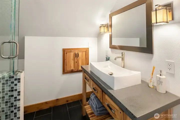 The private full bathroom servicing the space above the garage, beautifully styled with a modern vessel sink, custom concrete countertops, warm wood cabinetry, and a glass-enclosed shower with mosaic tile borders.