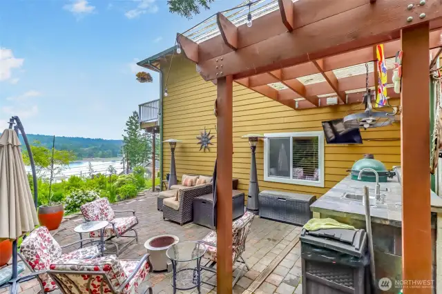 The expansive stone-paved patio features a complete outdoor kitchen, including a built-in BBQ, sink, and refrigerator, perfect for sophisticated alfresco dining. Relax or host guests on the comfortable sectional seating, or enjoy the ambiance created by the overhead pergola and outdoor heater.