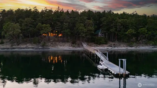 A striking sunset sky frames the long dock extending into calm waters, ideal for boaters, kayakers, or just soaking in the evening ambiance. The home sits tucked under the treeline, offering unmatched seclusion.