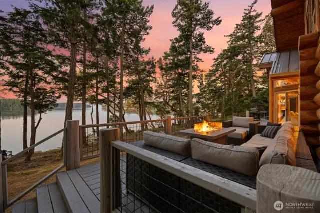 Enjoy serene evenings on the expansive deck surrounded by towering evergreens. A perfect space for gathering, all set against a stunning waterfront backdrop and glowing sunset skies.