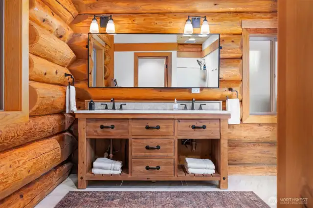 This primary, main level, full bath showcases rich log textures and a statement vanity with hand-forged hardware, blending luxury with rugged charm. Dual sinks and custom lighting complete the space.