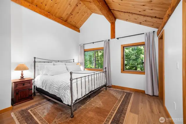 Vaulted cedar ceilings and large windows create a bright yet cozy retreat in the upper level guest room. The peaceful forest view, hardwood floors, and clean neutral palette offer a relaxing space to unwind.