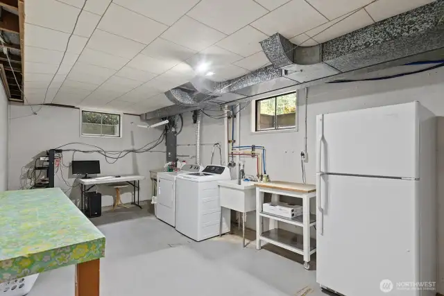 Spacious unfinished basement area with laundry and laundry folding table, extra fridge, wash basin and storage.