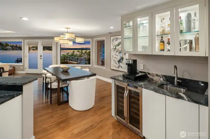 A fully appointed wet bar — marble countertop, wine refrigerator, undermount sink, and backlit glass cabinets stocked and ready — sets the stage for evenings that need no occasion. Beyond, the dining table glows beneath pendant light as Yarrow Bay fades into twilight through the windows. This is a home that knows how to host.