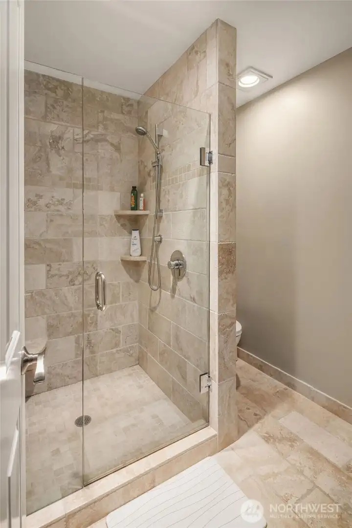 The en-suite bath delivers a spa-like experience in warm travertine — floor-to-ceiling stone tile wraps a generously sized walk-in shower enclosed in frameless glass, with a handheld shower, built-in corner shelving, and coordinating tile floors throughout. A quiet luxury that guests will not soon forget.